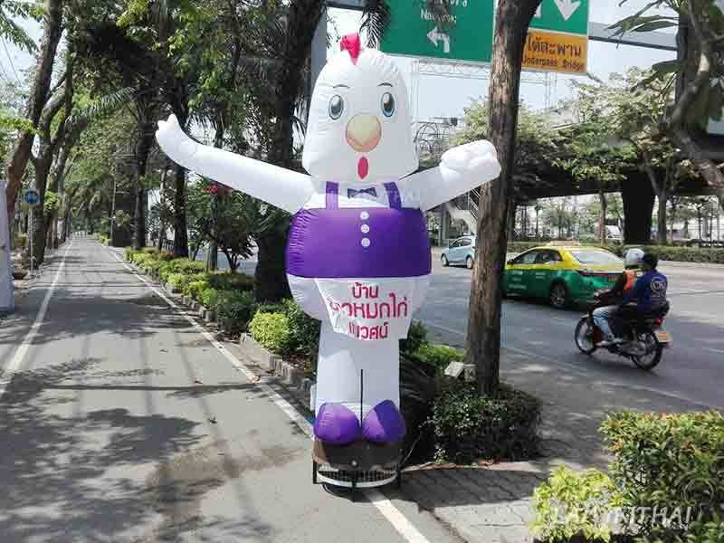 ตุ๊กตาโบกรูปไก่สีขาวผูกผ้ากันเปื้อนม่วง โฆษณาร้านอาหารริมถนนใหญ่